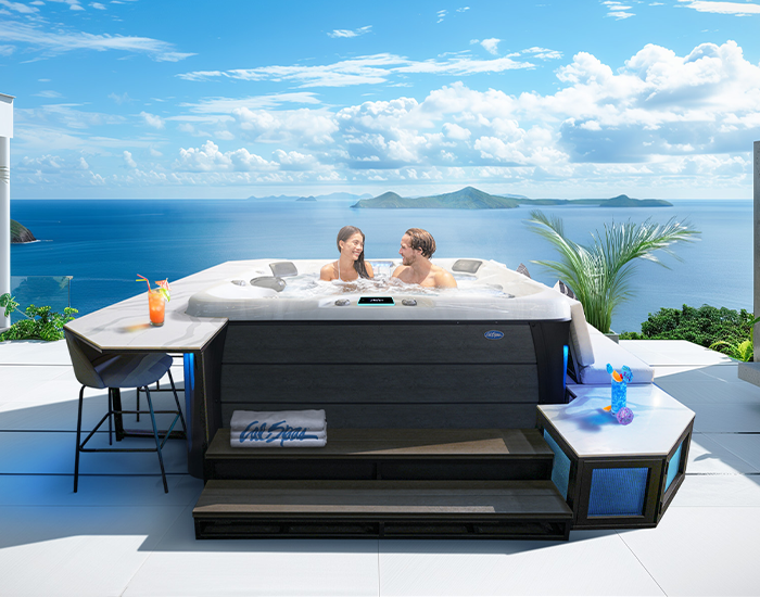 A couple smiling in a Cal Spas hot tub, with steps, bar and bench seat surrounds, on a patio overlooking an ocean