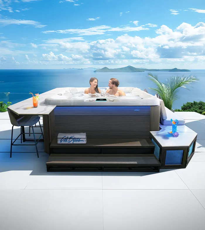 A couple smiling in a Cal Spas hot tub, with steps, bar and bench seat surrounds, on a patio overlooking an ocean